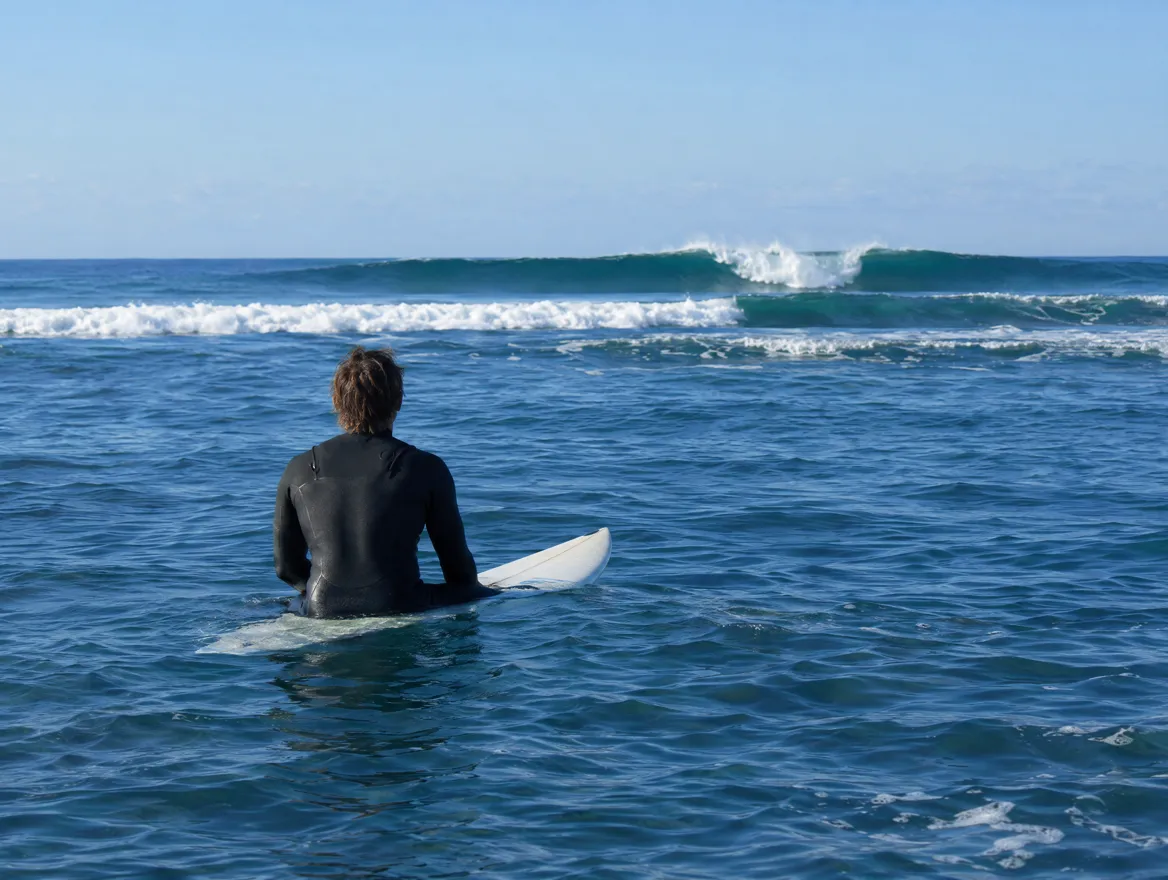 Surfer im Neoprenanzug sitzt von hinten gesehen auf seinem Brett im offenen Meer und beobachtet mehrere anrollende Wellen